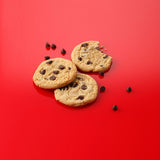 Three chocolate chip cookies arranged on a bright red background with scattered chocolate chips around them.