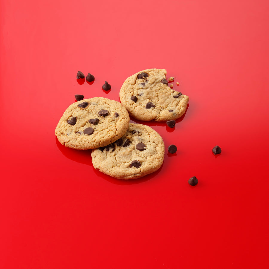 Three chocolate chip cookies arranged on a bright red background with scattered chocolate chips around them.
