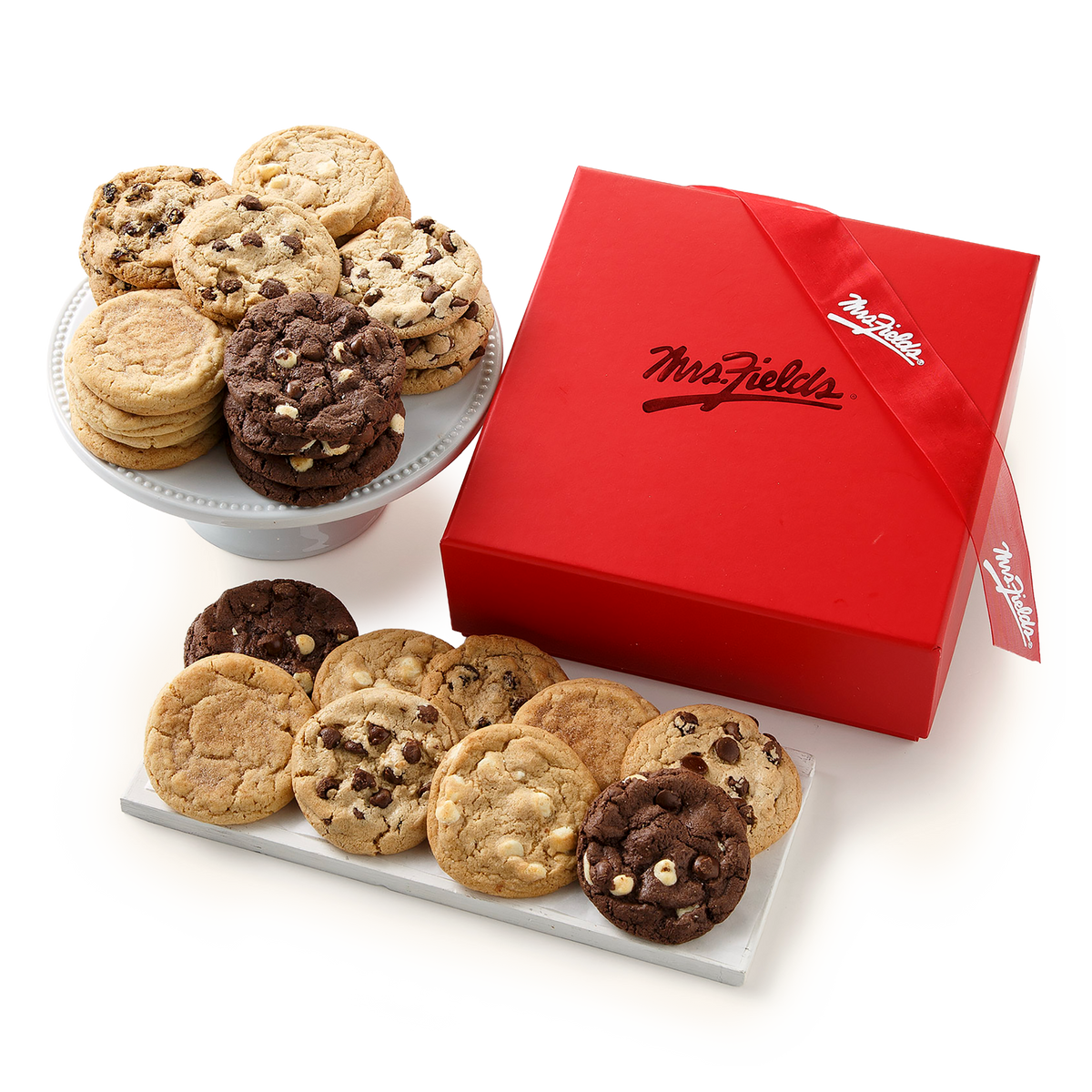 A gift assortment of cookies from Mrs. Fields, featuring various chocolate chip and oatmeal cookies displayed on a white stand and a white tray, alongside a red branded gift box with a ribbon.