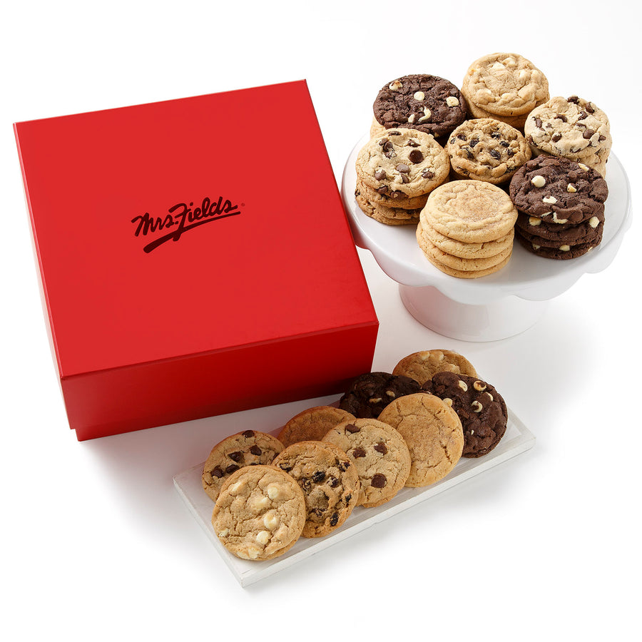 A large square red gift box with a shiny Mrs. Fields logo on it surrounded by an assortment of cookies on a blank white surface.