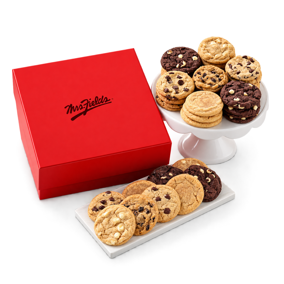 A red Mrs. Fields gift box with the brand name in black script, shown beside two white cake stands filled with assorted cookies, including chocolate chip cookies and chocolate chip cookies with white chocolate chips, on a clean white background.