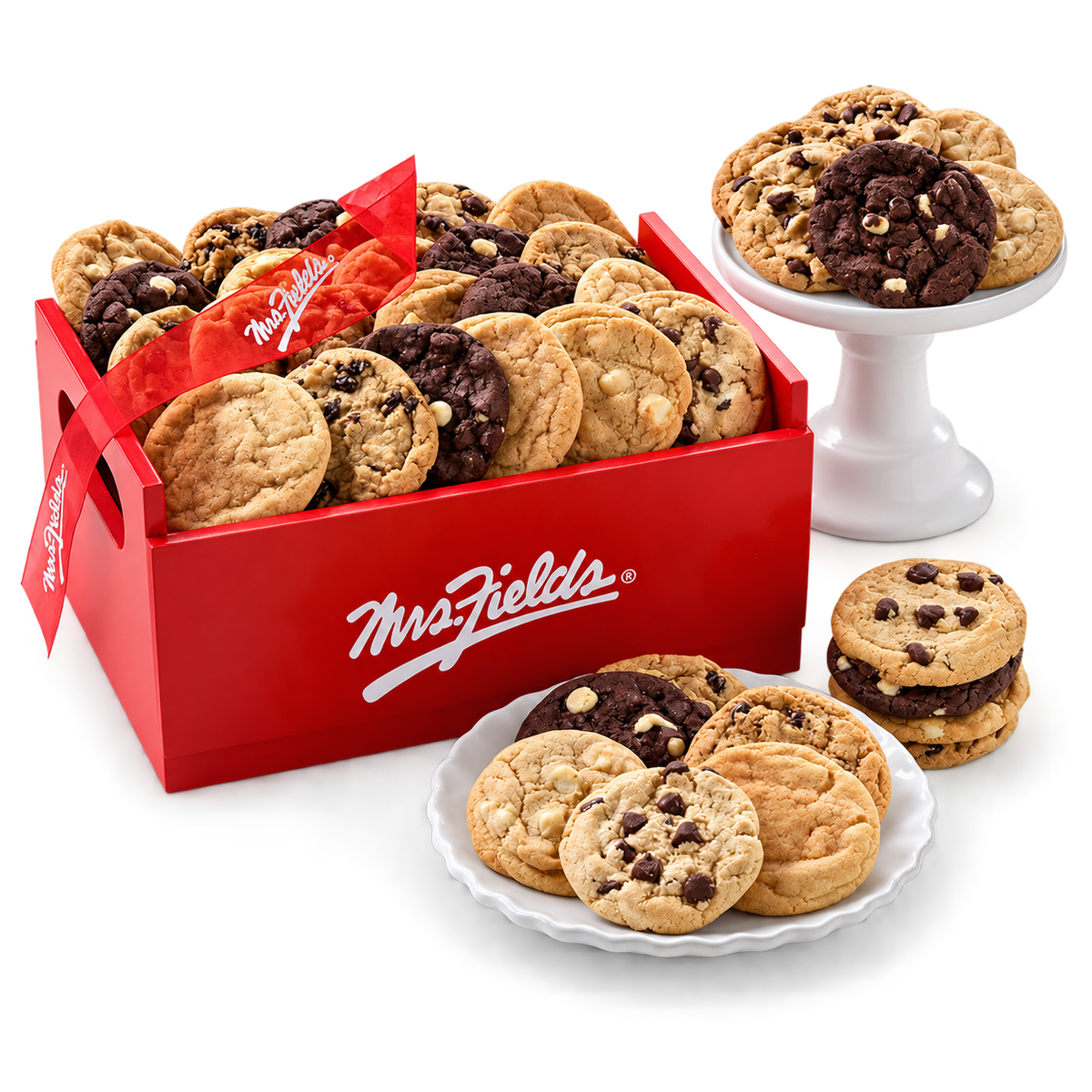 A red Mrs. Fields gift box filled with assorted cookies, with several cookies displayed on white plates and a white pedestal stand against a clean white background.