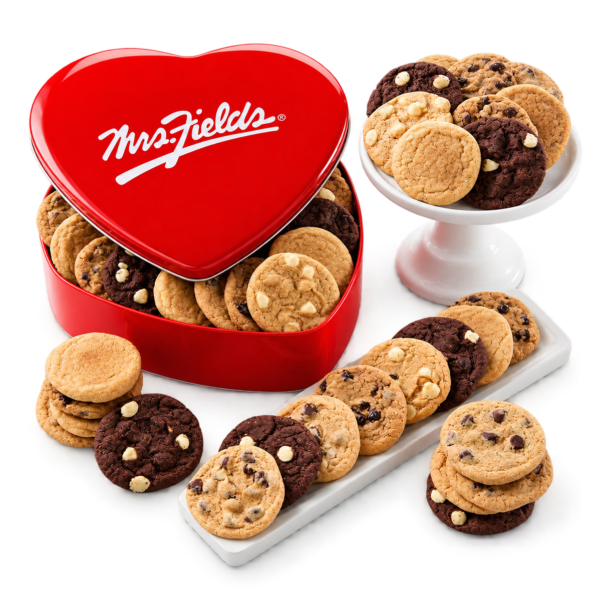 A heart-shaped red Mrs. Fields cookie tin filled with assorted cookies, styled on a white background with additional cookies arranged on white platters and stands.