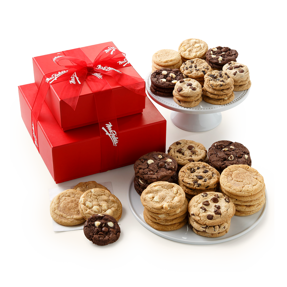 A gift set of assorted cookies displayed on white plates and a cake stand alongside two stacked red gift boxes tied with a red ribbon. The cookies include chocolate chip, oatmeal cookie, and dark chocolate varieties.