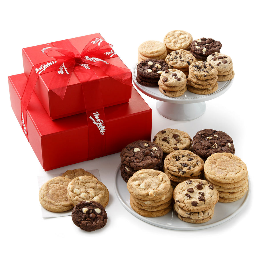 A two-tiered red tower surrounded by an assortment of both big and small cookies with a red Mrs. Fields ribbon on it on a blank white surface.