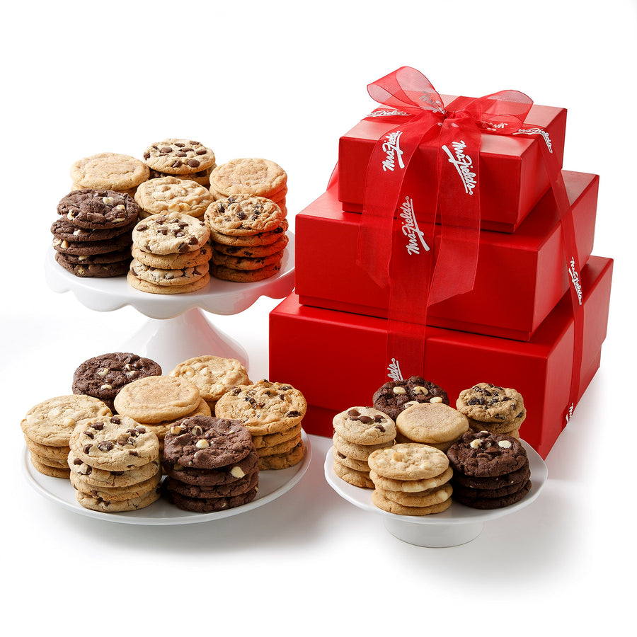 A three-tiered red gift tower composed of three red gift boxes and tied with a red Mrs. Fields ribbon surrounded by a large assortment of both big and small cookies on a blank white surface.