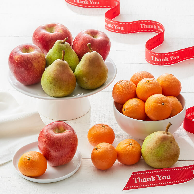 A styled fruit arrangement on white plates and a bowl, including red apples, green and yellow pears, and oranges, with red ribbon accents printed with the words 'Thank You'.