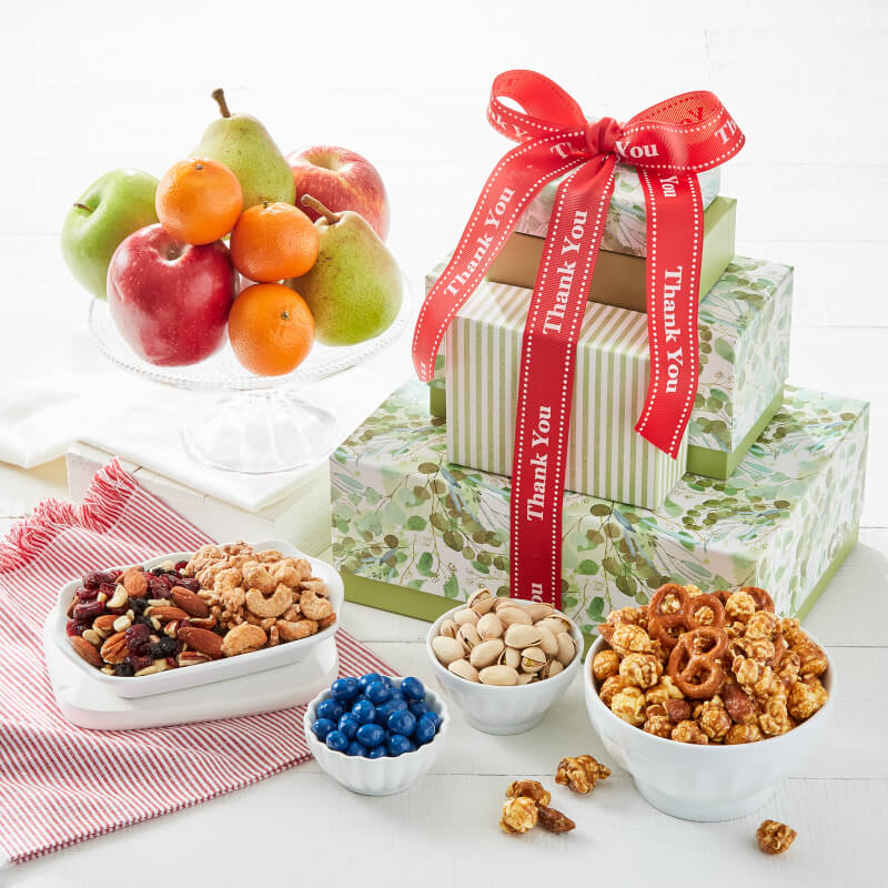 A gift basket arrangement on a white surface featuring three stacked gift boxes wrapped in green botanical paper and tied with a red ribbon printed with 'Thank You,' alongside bowls of snacks and a fruit stand with apples, oranges, and pears.