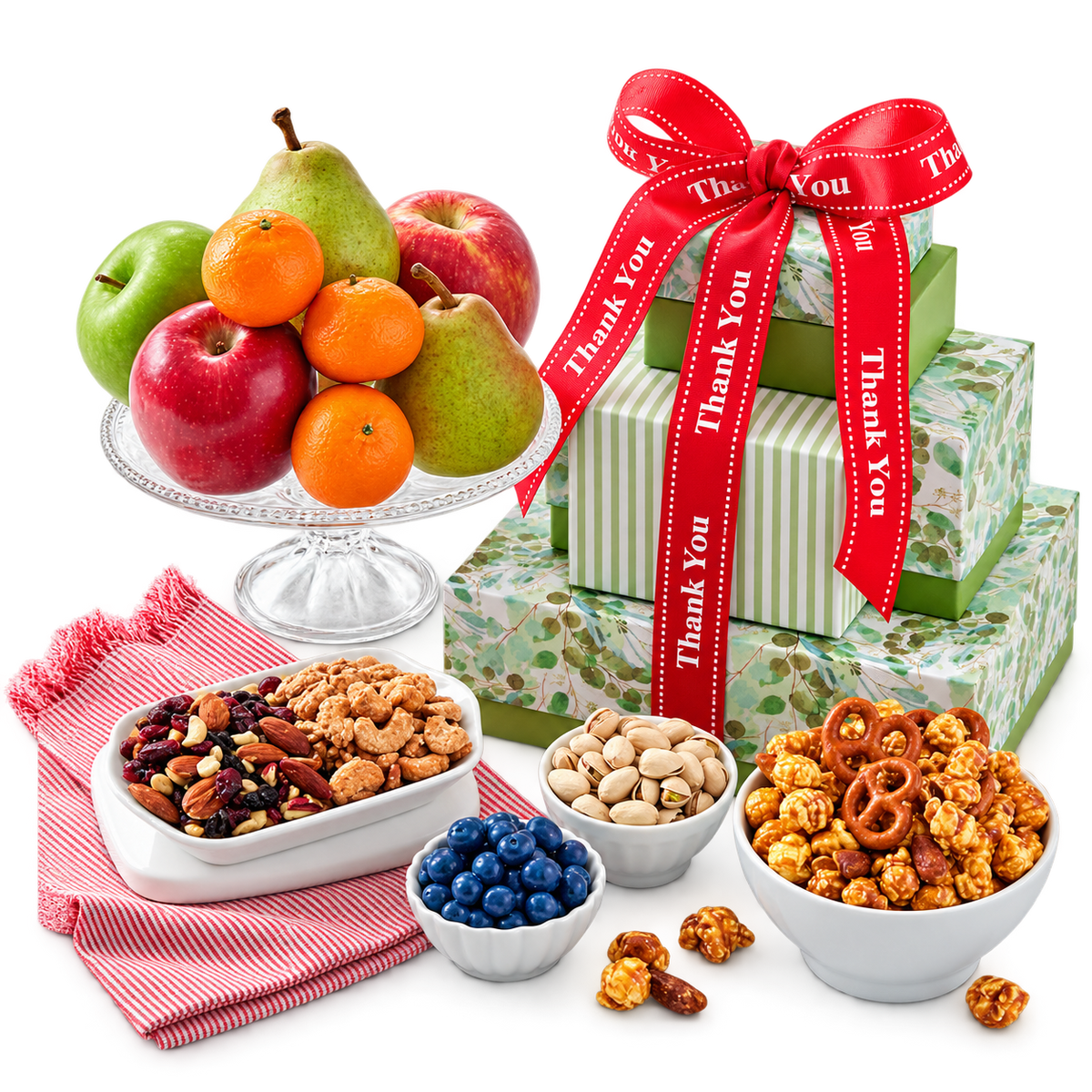 A gift basket arrangement on a white surface featuring three stacked gift boxes wrapped in green botanical paper and tied with a red ribbon printed with 'Thank You,' alongside bowls of snacks and a fruit stand with apples, oranges, and pears.