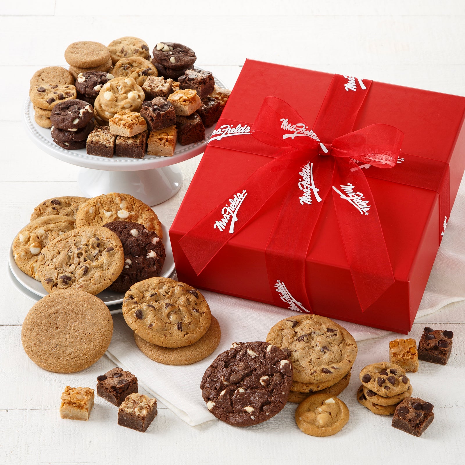 A red gift box tied with a red Mrs. Fields ribbon and surrounded by an assortment of Nibblers®, original cookies, and brownie bites.