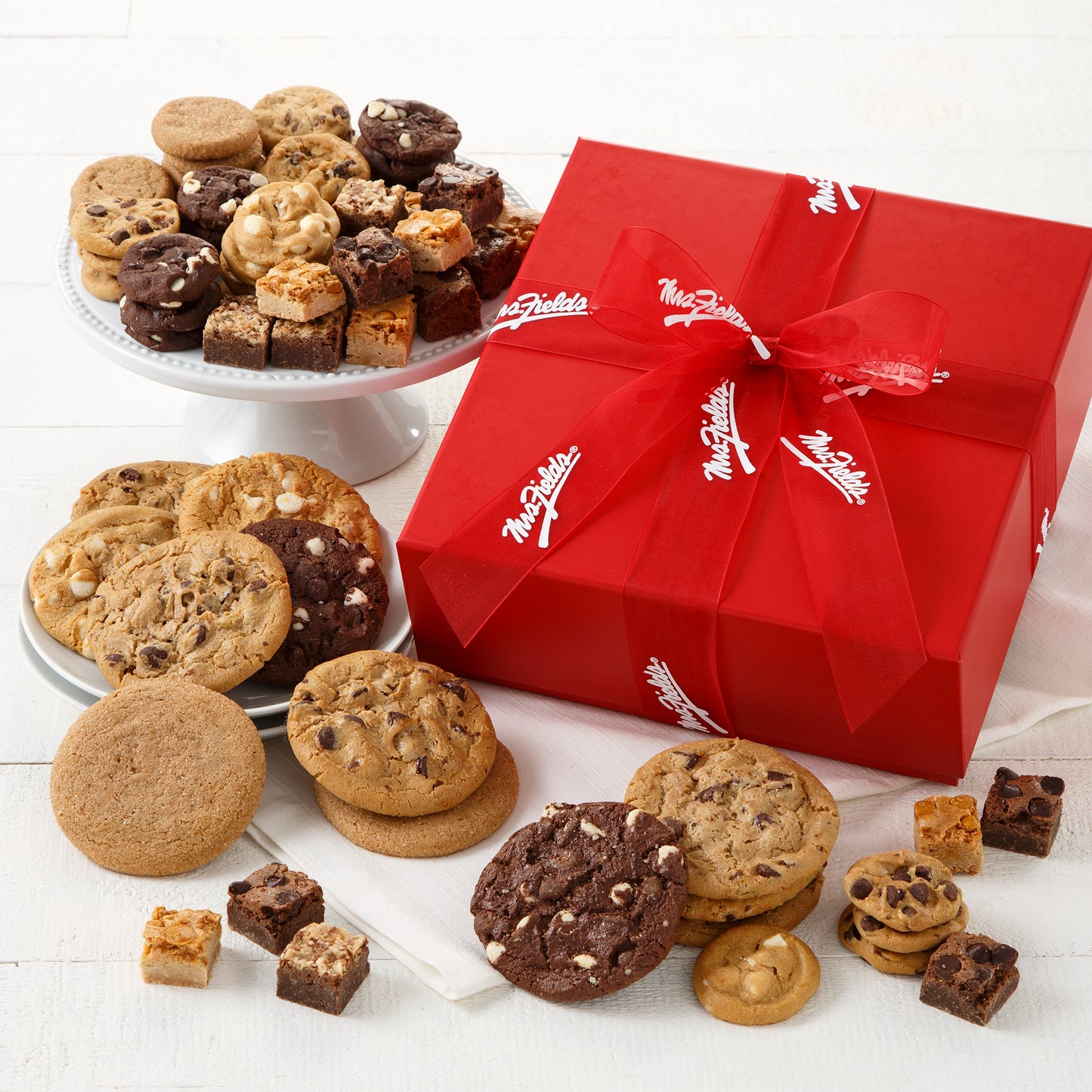 A red gift box tied with a red Mrs. Fields ribbon and surrounded by an assortment of Nibblers®, original cookies, and brownie bites