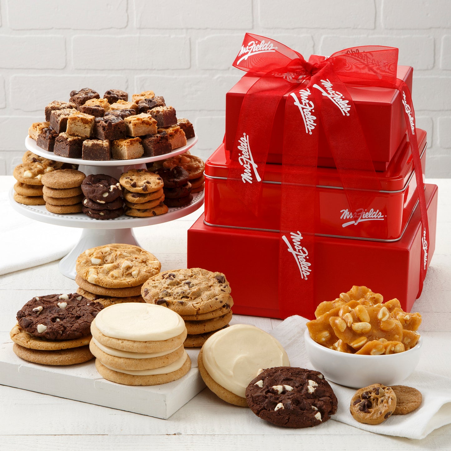 A three tier red tower tied with a Mrs. Fields ribbon and surrounded by an assortment of Nibblers®, original cookies, brownie bites, frosted cookies, and peanut brittle