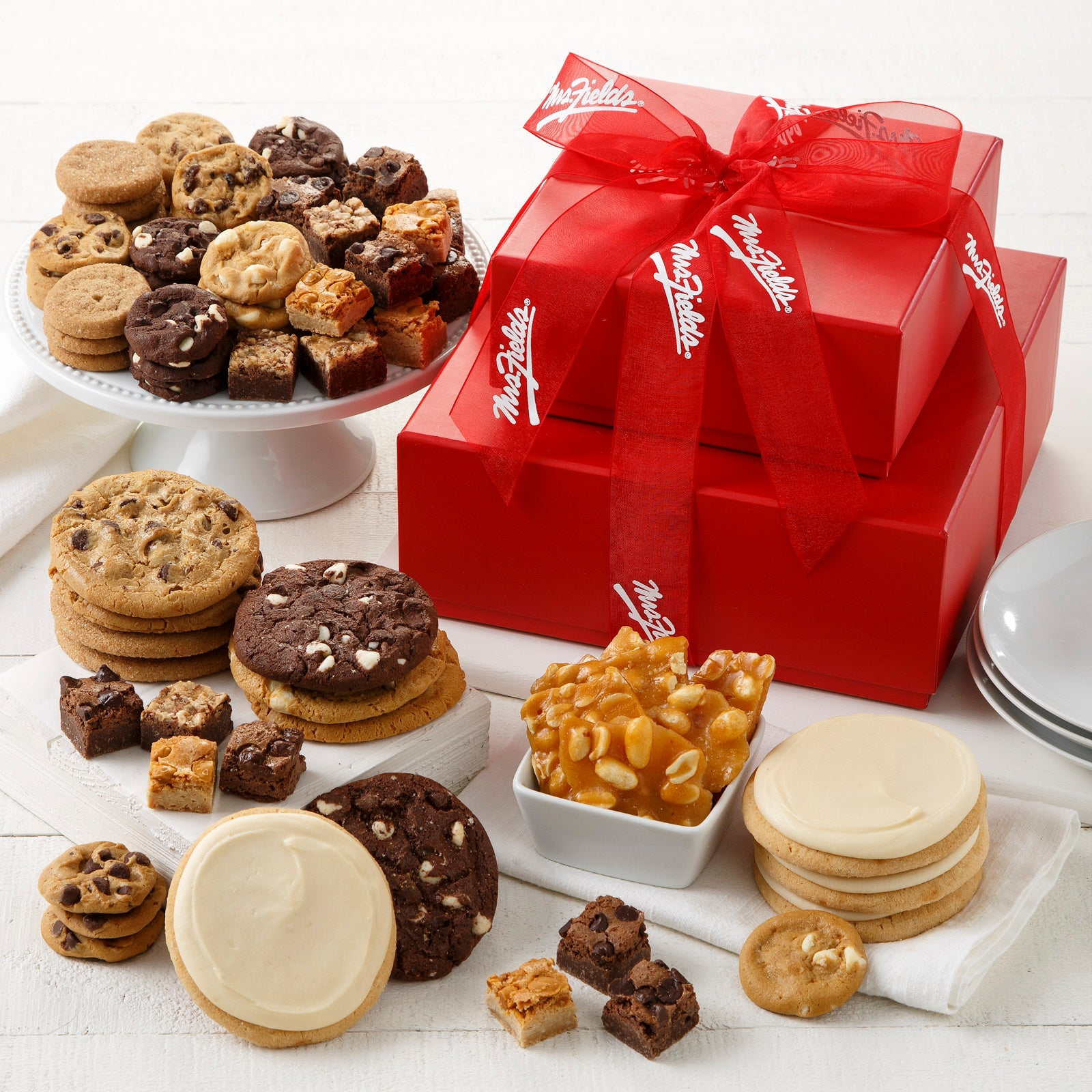 A two tier red box tower tied with a red Mrs. Fields ribbon and surrounded by an assortment of Nibblers®, original cookies, frosted cookies, brownie bites, and peanut brittle