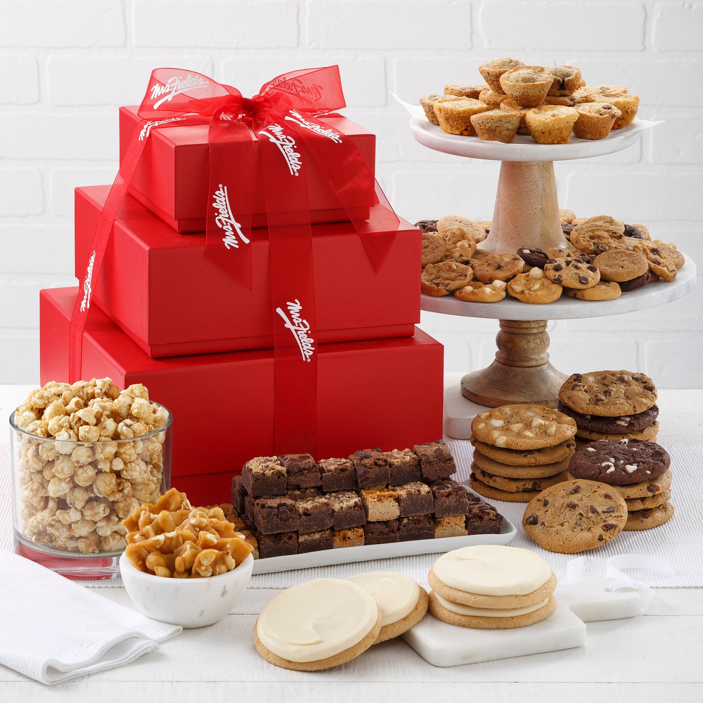 A three tier red tower tied with a Mrs. Fields ribbon and surrounded by an assortment of Nibblers®, original cookies, brownie bites, frosted cookies, popcorn, and peanut brittle