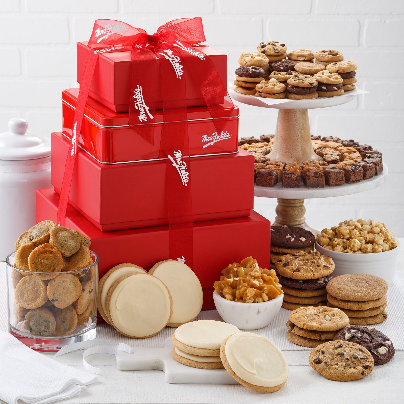 A four tier red tower tied with Mrs Fields ribbon and surrounded by an assortment of Nibblers®, original cookies, brownie bites, frosted cookies, muffins, popcorn, and peanut brittle.