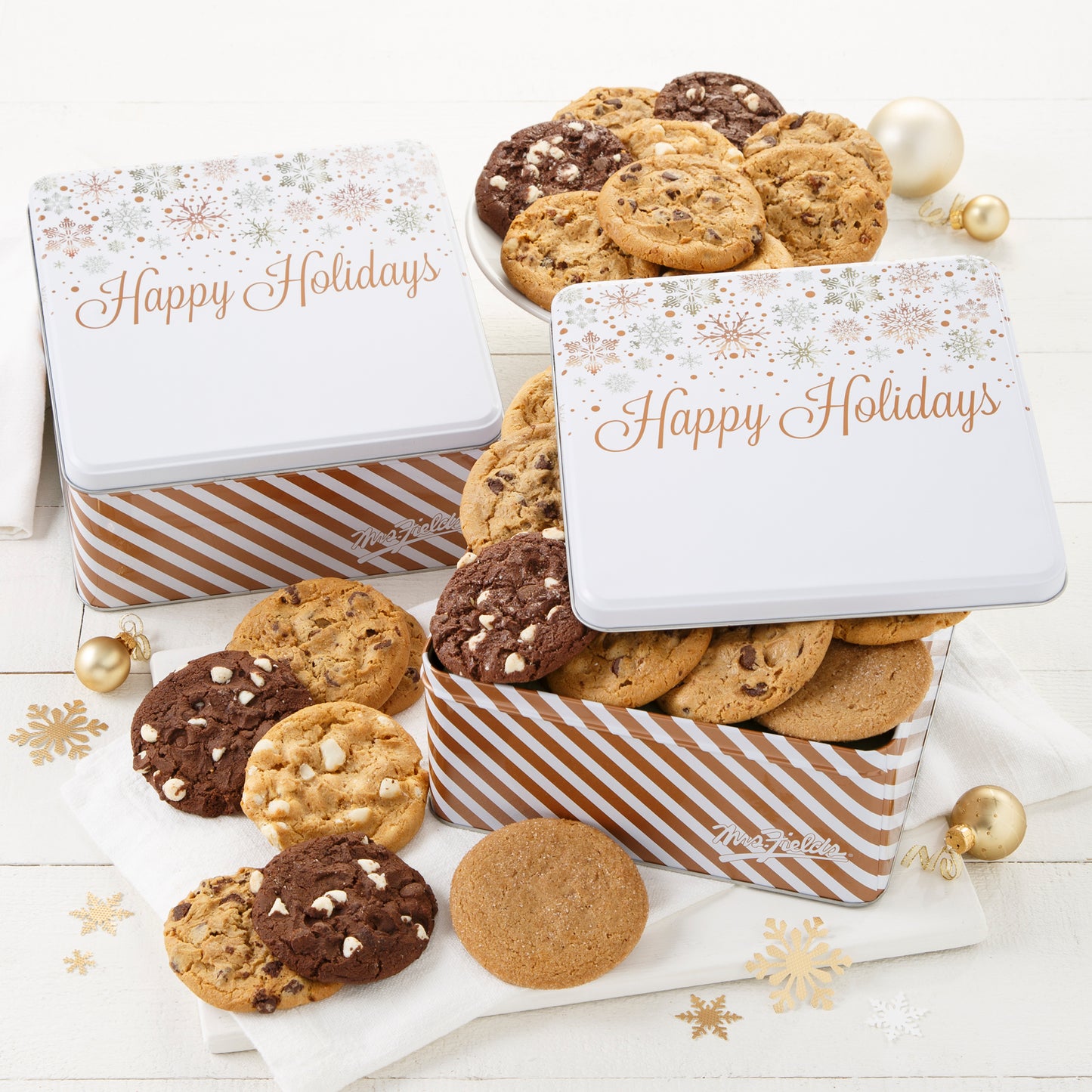 Two Happy Holidays gift tins decorated with silver and white snowflakes on a white top and filled with an assortment of original cookies.