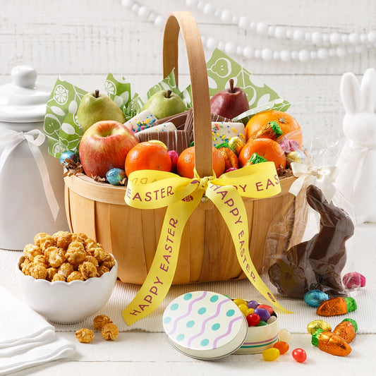 Wooden basket filled with an assortment of treats including fresh fruits, chocolates, jelly beans, caramel corn, and a chocolate bunny all on a white surface with bunny figurines.
