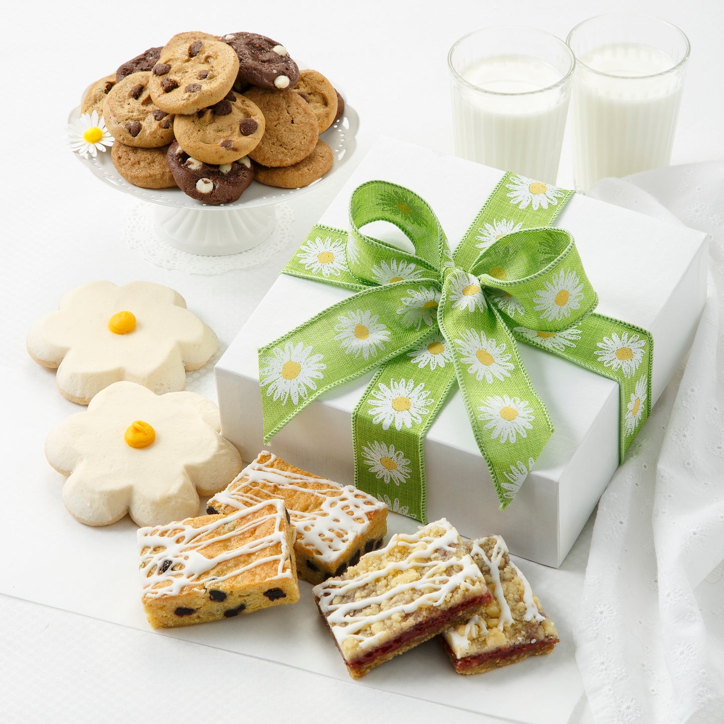 A white linen box tied with a green daisy themed ribbon. Surrounding this gift box is an assortment of Nibblers®, two frosted white flower cookies and an assortment of fruit bars