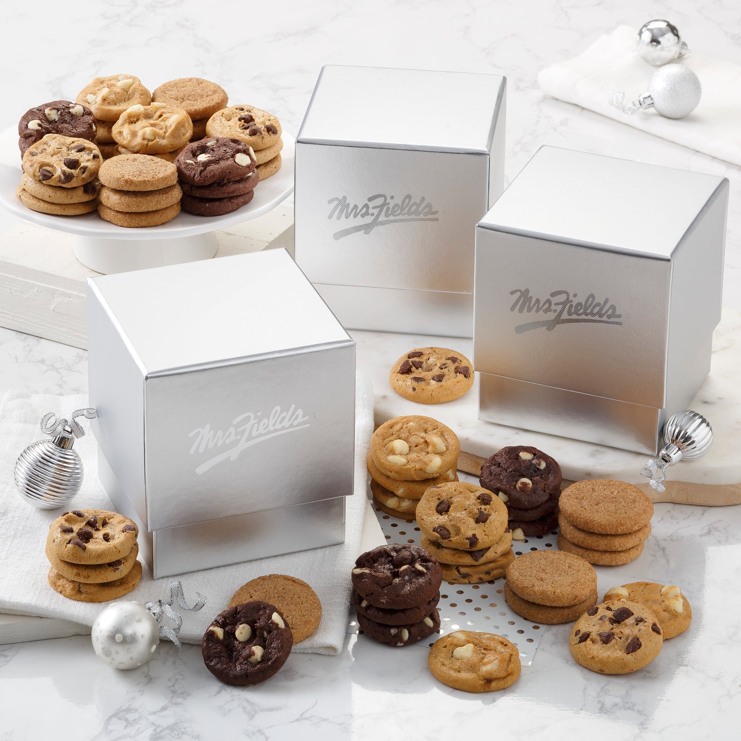 Three silver Mrs. Fields mini gift boxes surrounded by an assortment of Nibblers®
