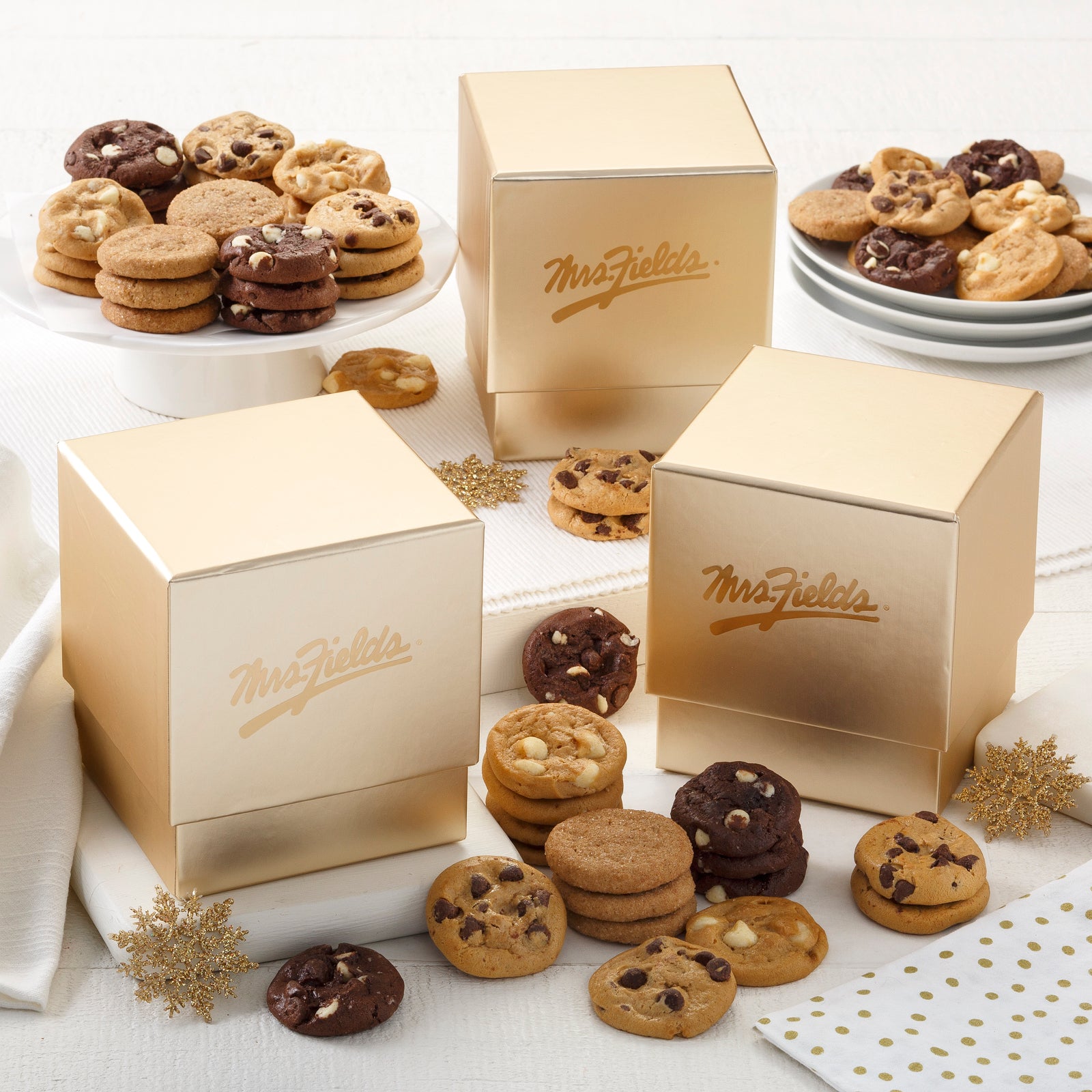 Three gold Mrs. Fields mini gift boxes surrounded by an assortment of Nibblers®