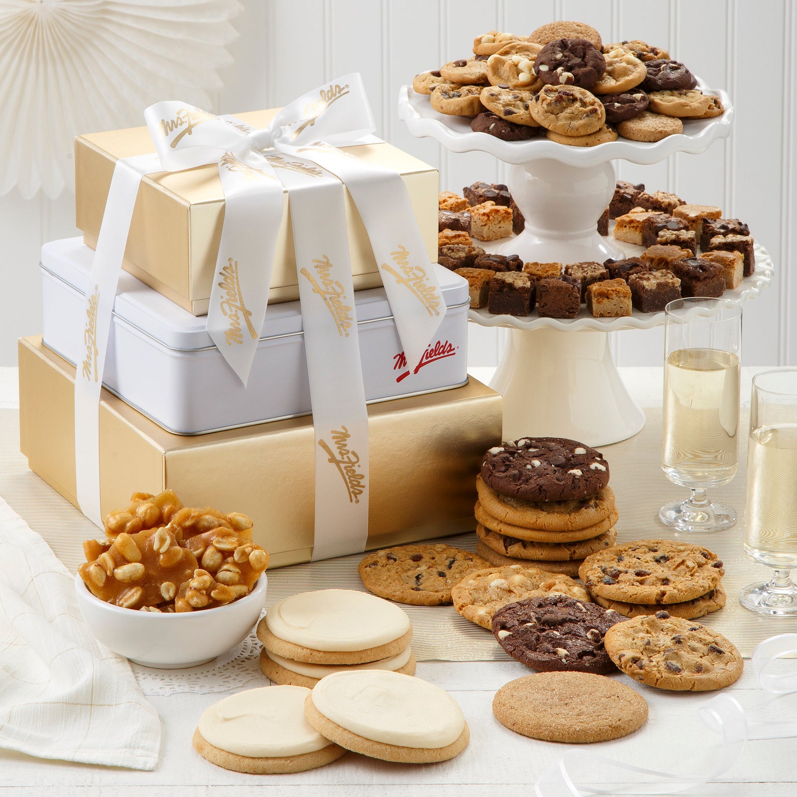 A three-tier gold and white tower tied with a white and gold Mrs. Fields ribbon and surrounded by an assortment of Nibblers®, original cookies, four frosted cookies, peanut brittle. 
