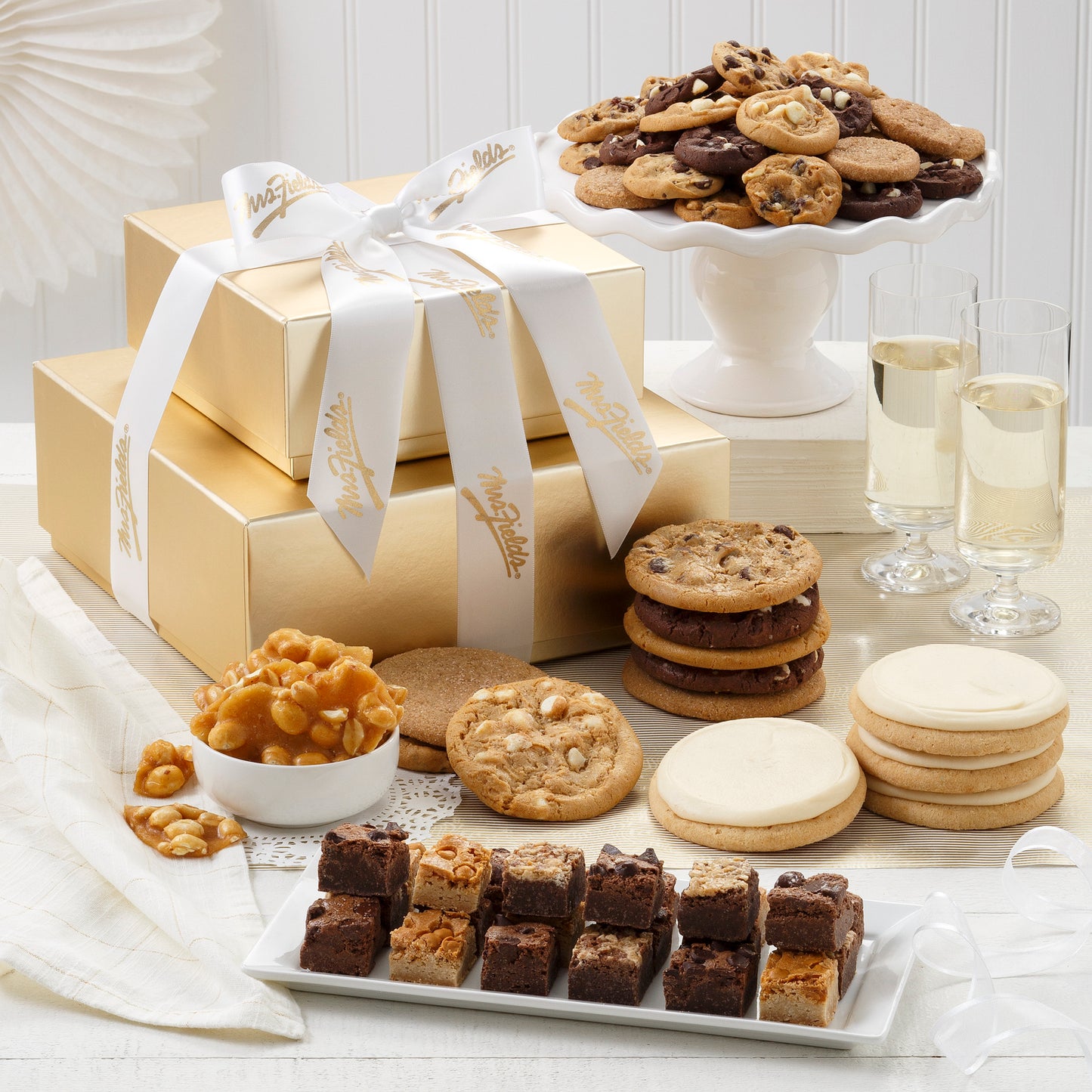 A two-tier gold box tower tied with a white and gold Mrs. Fields ribbon and surrounded by an assortment of Nibblers®, original cookies, four frosted cookies, and peanut brittle.