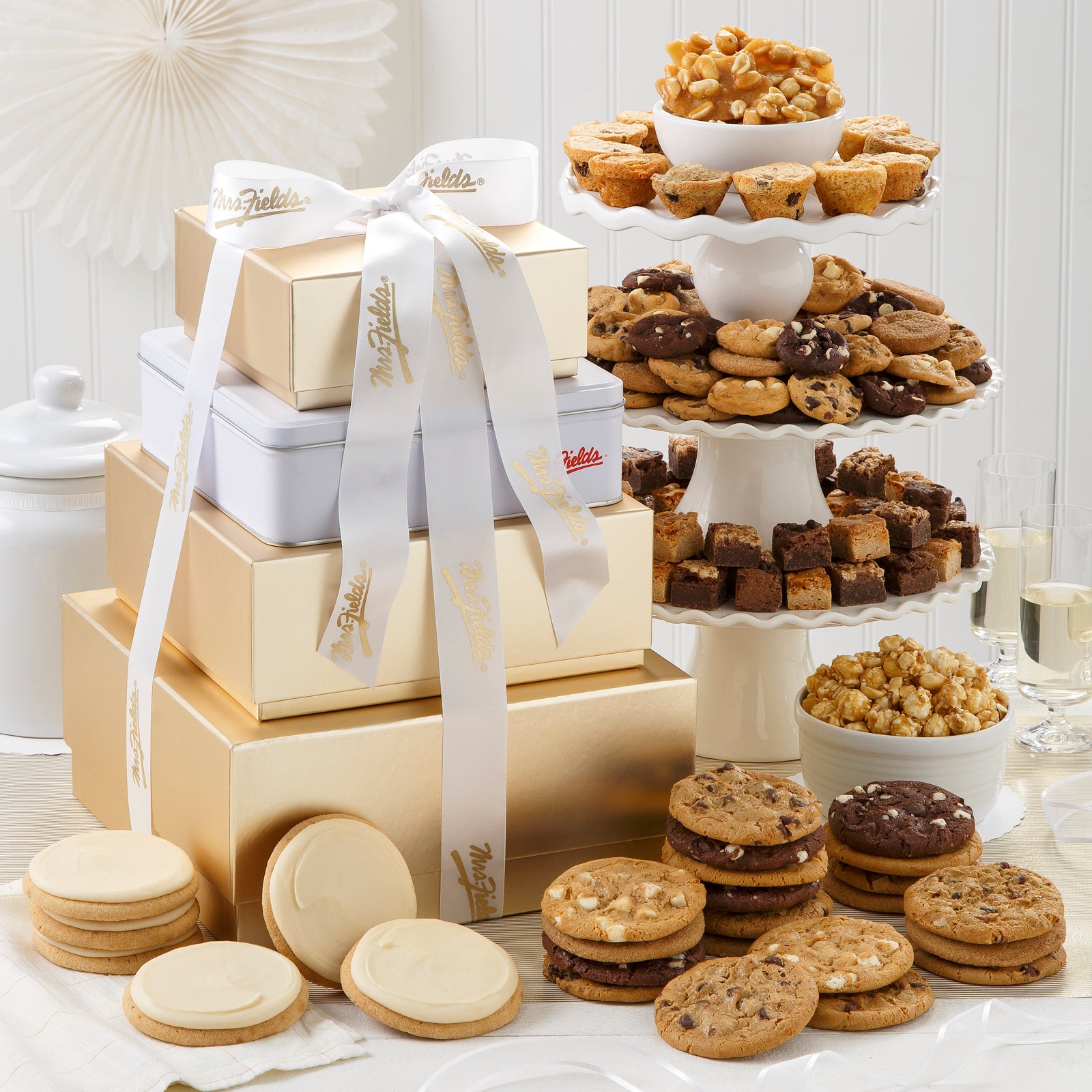 A four-tier gold and white tower tied with a white and gold Mrs. Fields ribbon and surrounded by an assortment of 75 Nibblers®, brownie bites, original cookies, muffins, 6 frosted cookies, peanut brittle, and popcorn.