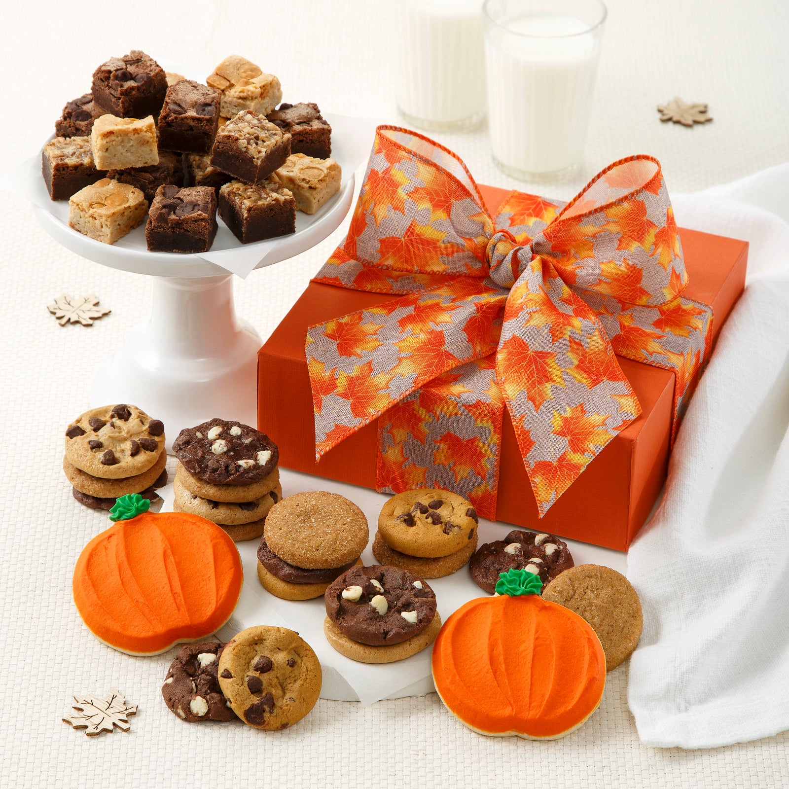 Orange gift box tied with fall themed orange ribbon with an assortment of Nibblers®, brown bites, and two frosted pumpkin cookies surrounding it