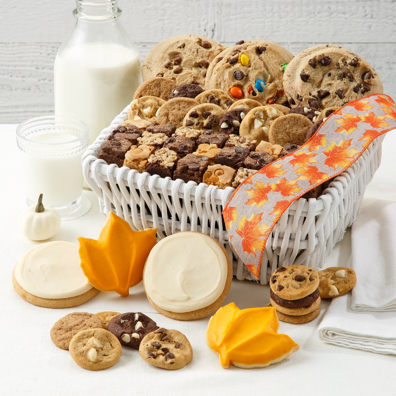 Small white basket filled with mega cookies, Nibblers®, and brownie bites. In front of basket is an assortment of Nibblers®, two round frosted cookies and two leaf cookies.