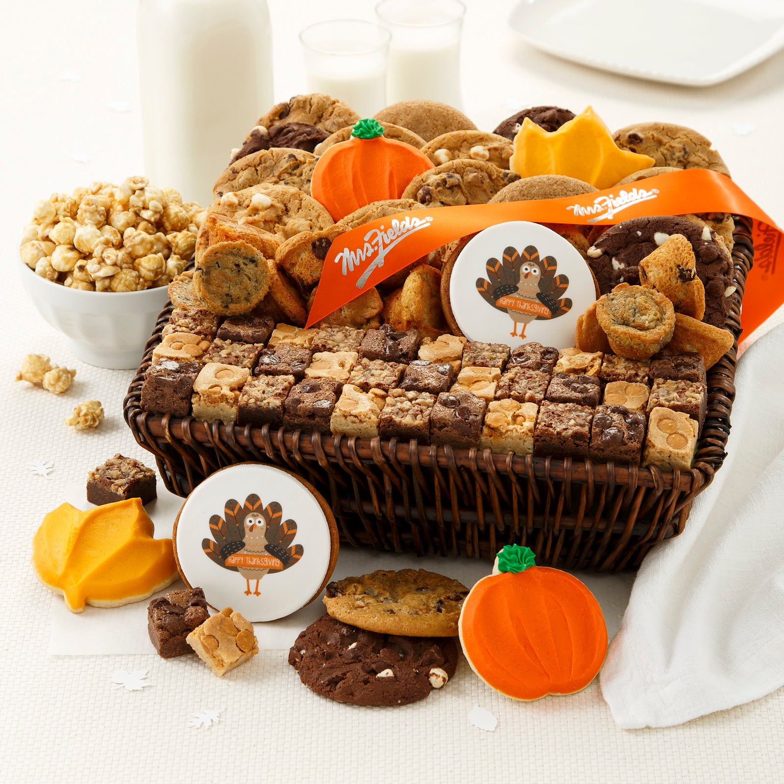 A large brown basket filled with an assortment of original cookies, brownie bites, muffins, frosted cookies, and a logo cookie. Outside of the basket is a bowl of popcorn with an assortment of frosted cookies, brownie bites and original cookies