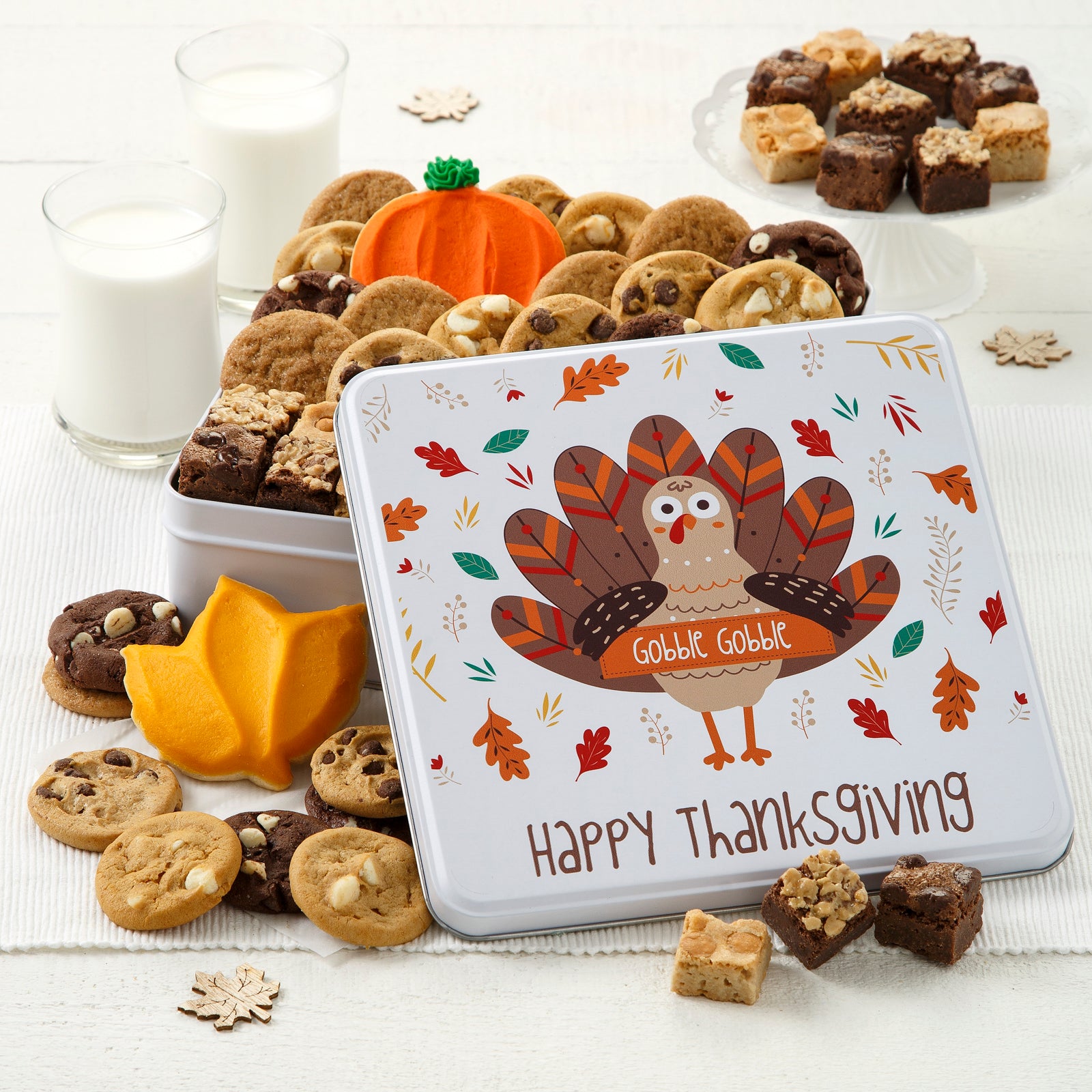 A Happy Thanksgiving them tin with a festive turkey on the lid. This tin is filled with an assortment of Nibblers®, brownie bites and a frosted pumpkin cookie. Surrounding the tin is an assortment of Nibblers®, brownie bites, and a frosted leaf cookie.