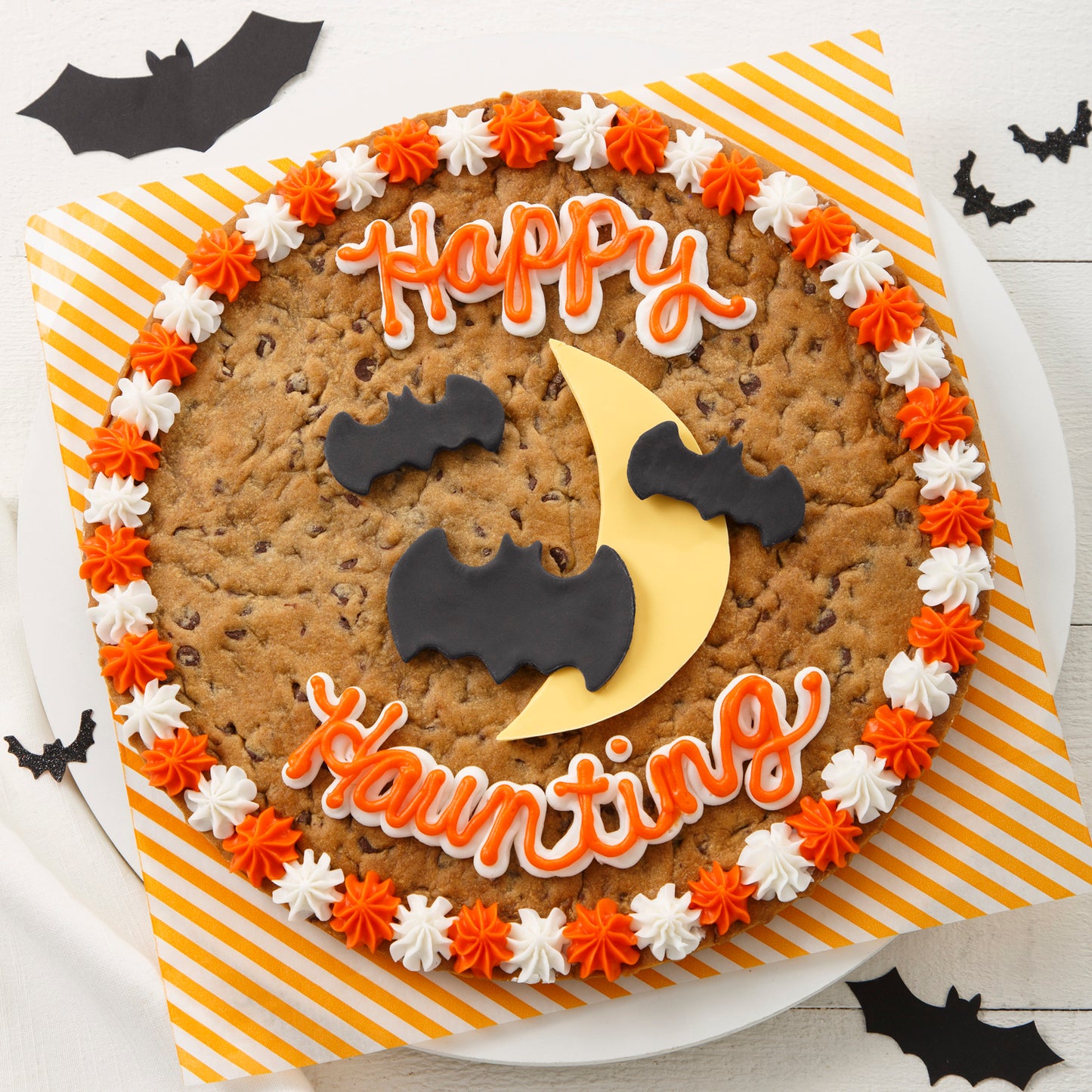 Halloween themed cookie cake with a Happy Haunting sentiment written in orange and white frosting. In the middle is a half moon with bats in fondant frosting. 