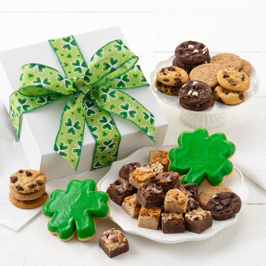 A white linen gift box tied with a green St. Patrick's Day ribbon and surrounded by an assortment of nibblers, brownie bites, and two frosted shamrock cookies