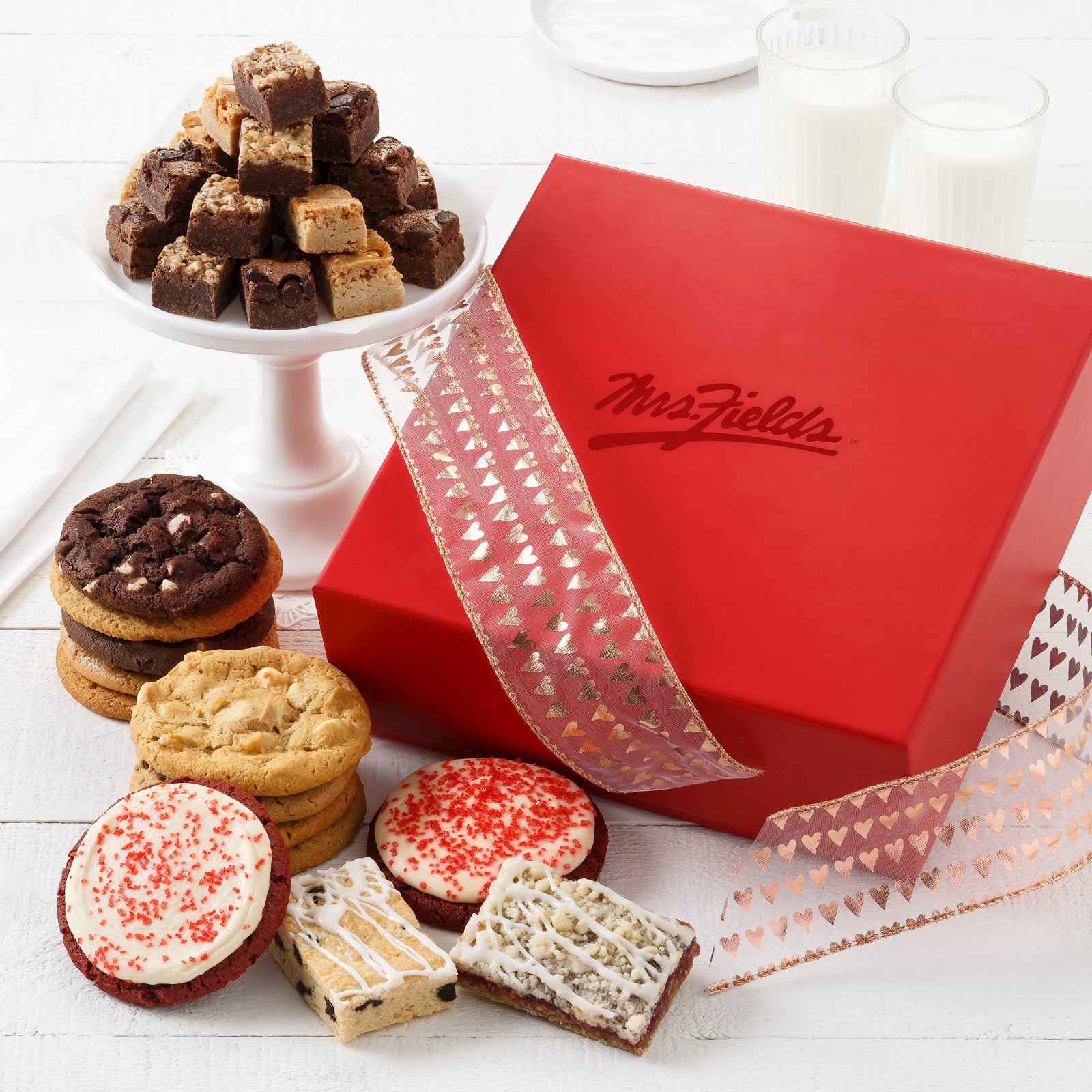 A red Mrs. Fields gift box with a heart ribbon and surrounded by an assortment of original cookies, brownie bites, fruit bars, and two frosted red velvet cookies with sprinkles