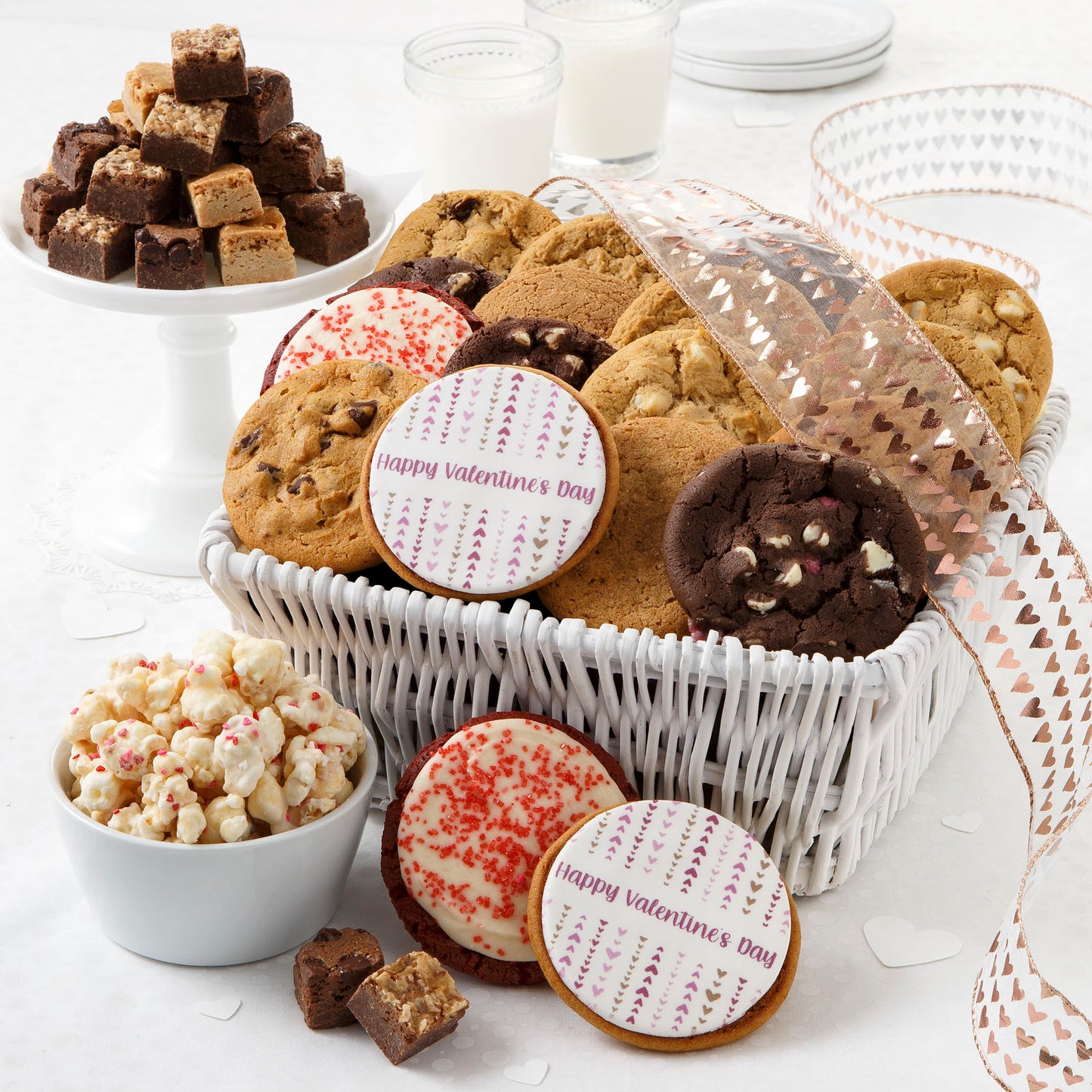 A white basket tied with a heart ribbon and filled with an assortment of original cookies, brownie bites, two frosted red velvet cookies, two logo cookies, and popcorn