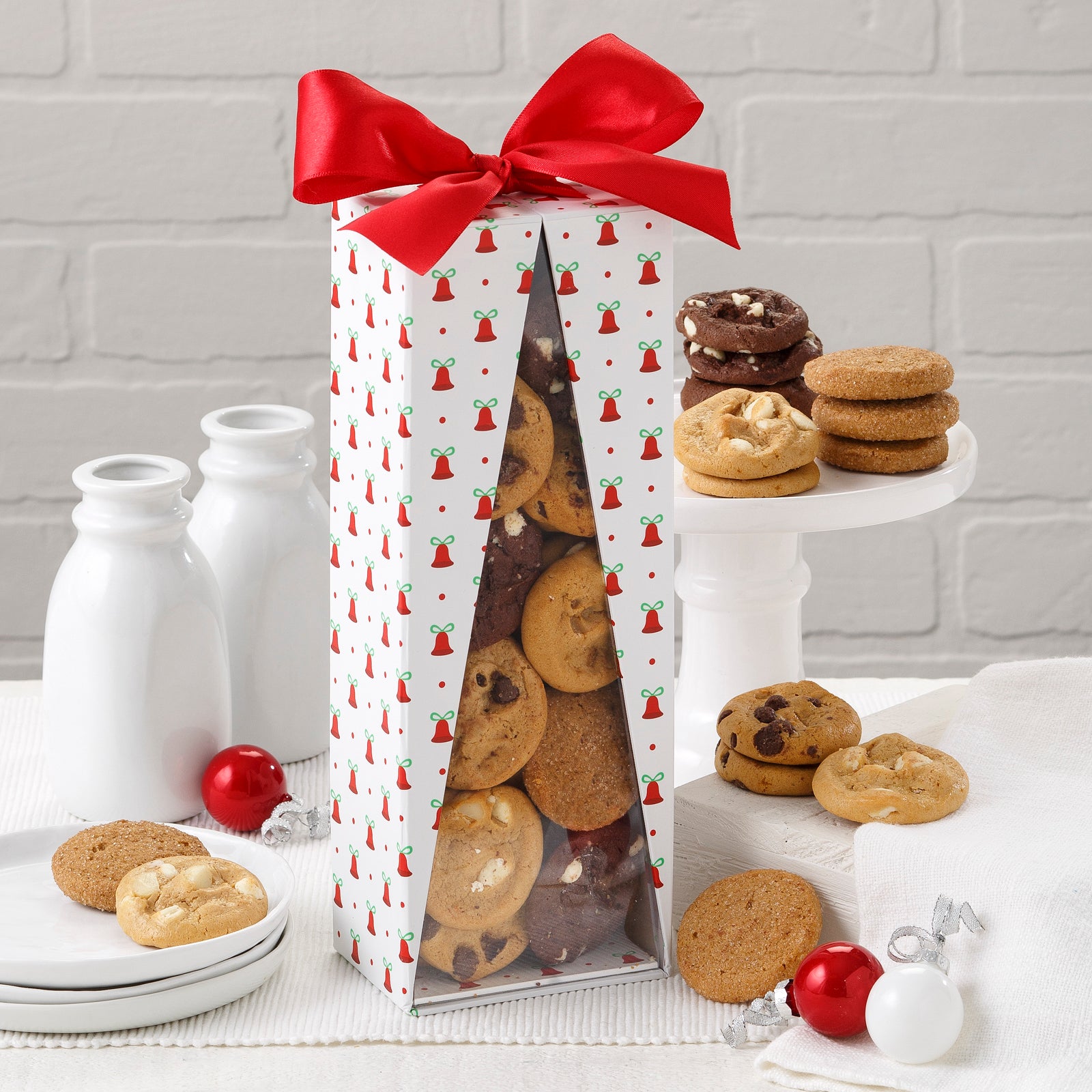 A holiday-themed gift box covered in bells with a red ribbon on top filled with an assortment of Nibblers® Bite-Sized Cookies.