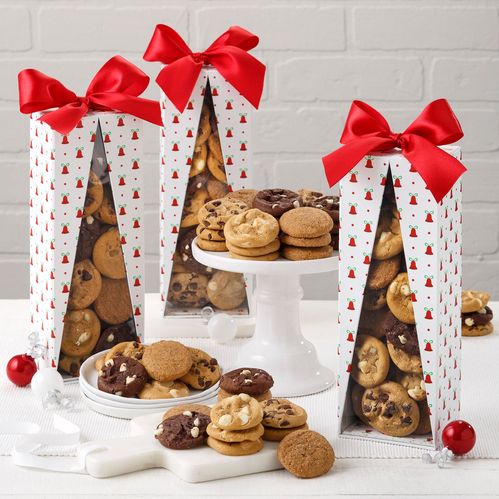 A group of holiday-themed gift boxes covered in bells with a red ribbon on top filled with an assortment of Nibblers® Bite-Sized Cookies.