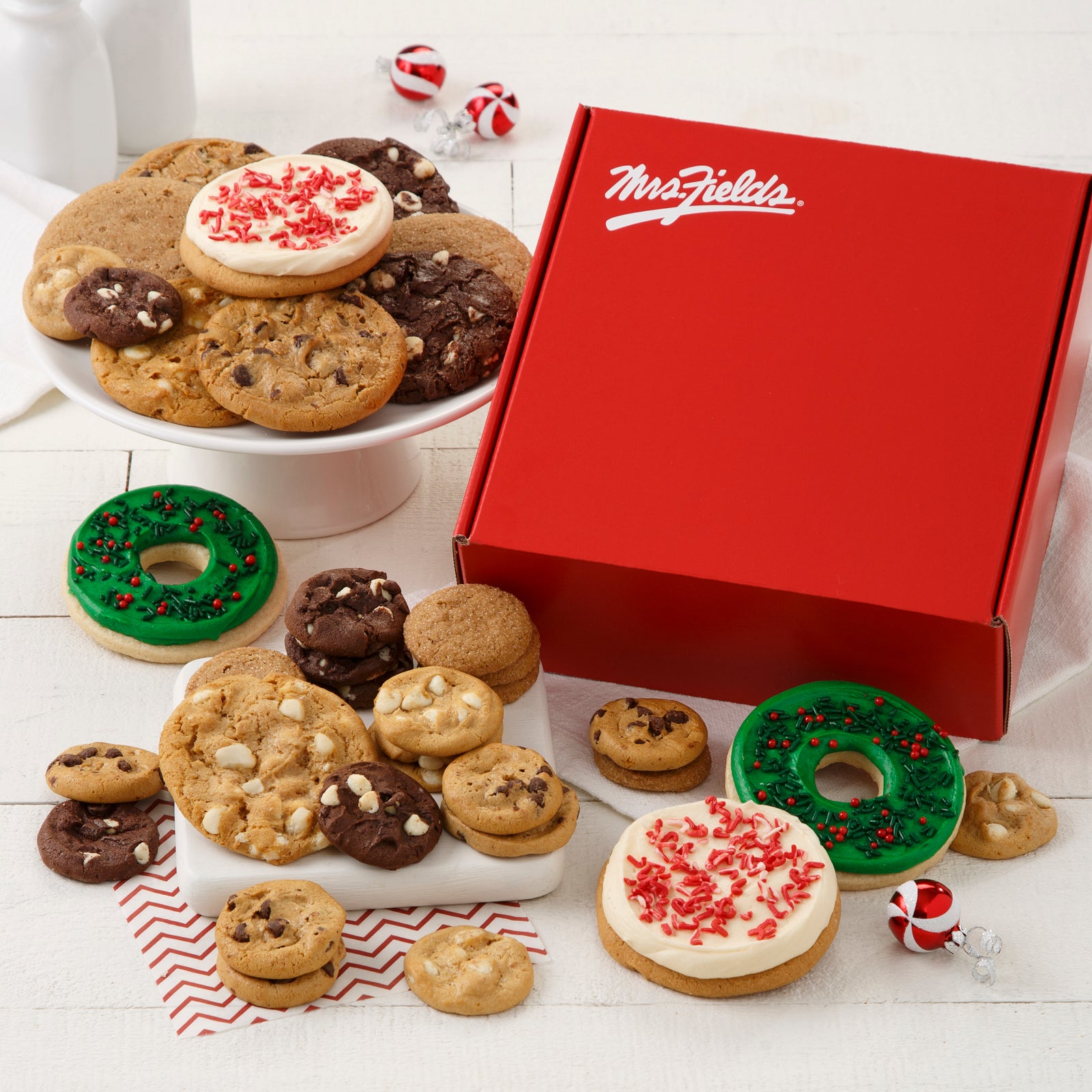 A classic red Mrs. Fields gift box with an assortment of original cookies, Nibblers®, two frosted wreaths with sprinkles, and two peppermint round cookies with sprinkles.