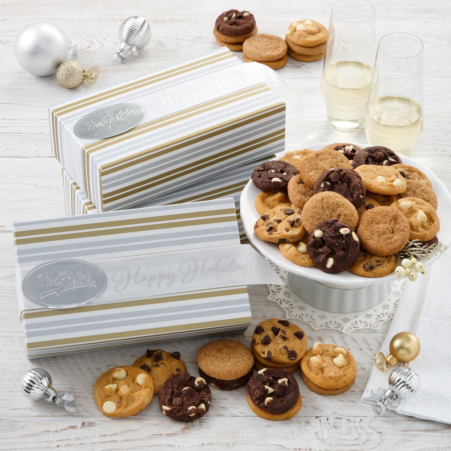 Three Happy Holidays gift boxes that are decorated with white, gold, and silver stripes and surrounded with Nibblers®
