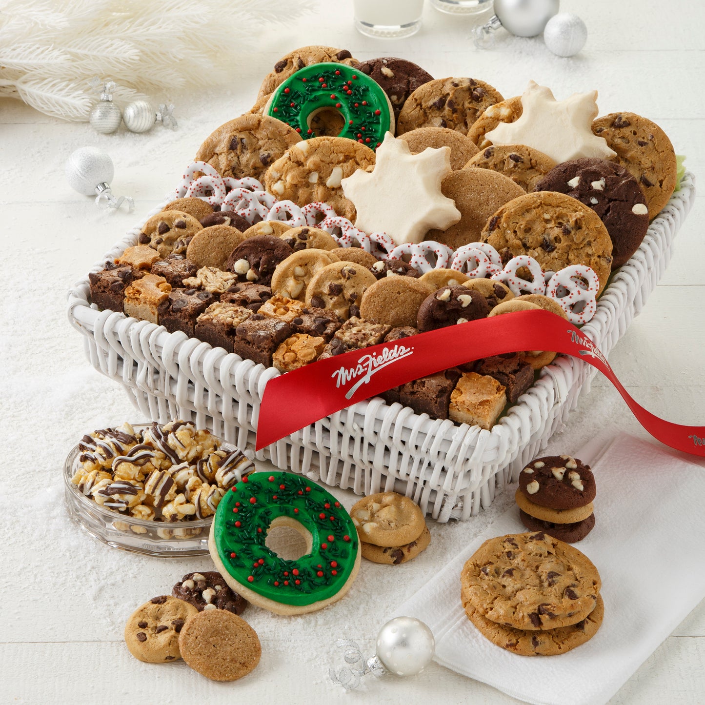 A white basket filled with an assortment of original cookies, Nibblers®, brownie bites, yogurt-covered pretzels, two frosted snowflake cookies, two frosted wreath cookies with sprinkles, and chocolate drizzled popcorn and decorated with a red Mrs. Fields' ribbon.