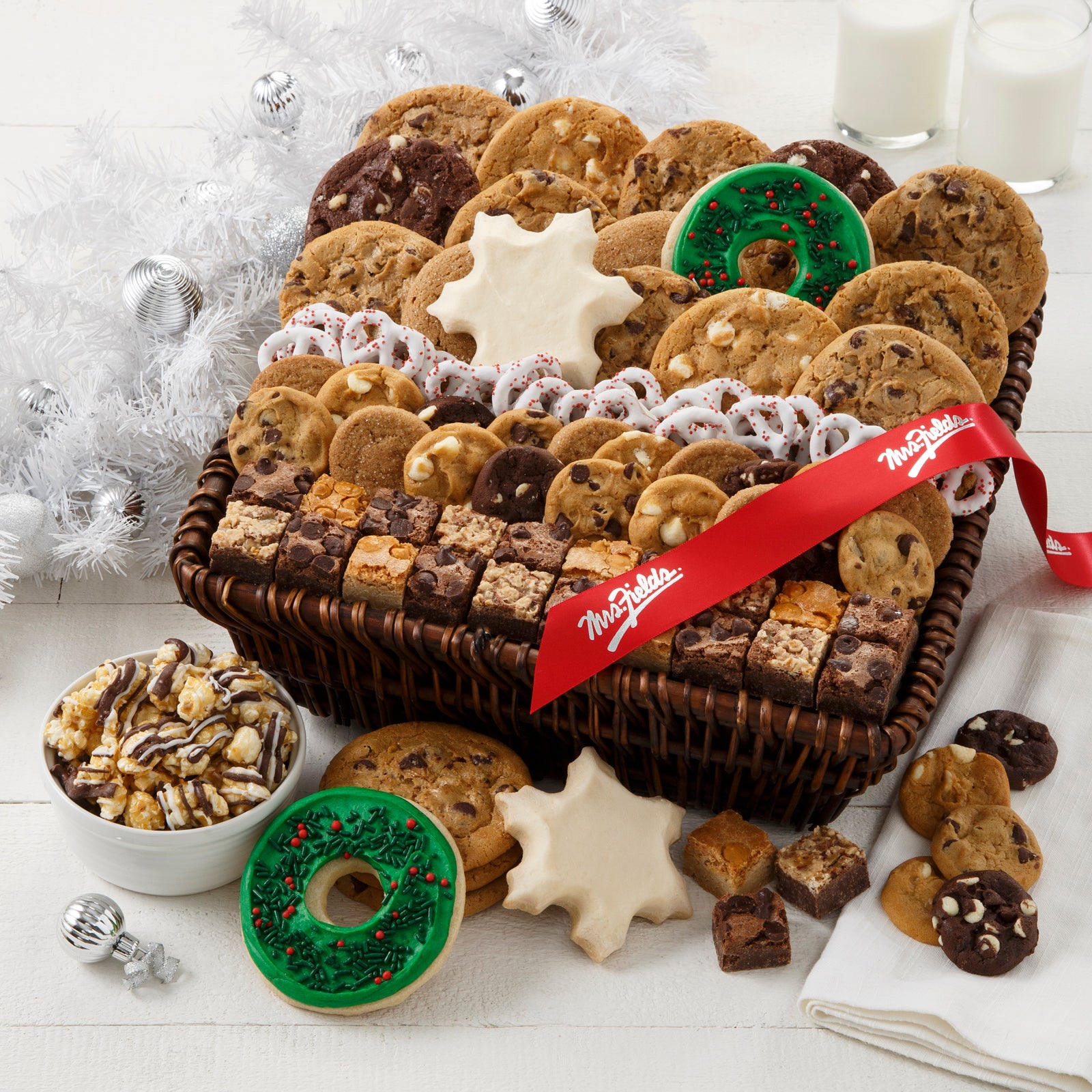 A brown basket filled with an assortment of original cookies, Nibblers®, brownie bites, yogurt-covered pretzels, two frosted snowflake cookies, two frosted wreath cookies with sprinkles, and chocolate drizzled popcorn and decorated with a red Mrs. Fields' ribbon.