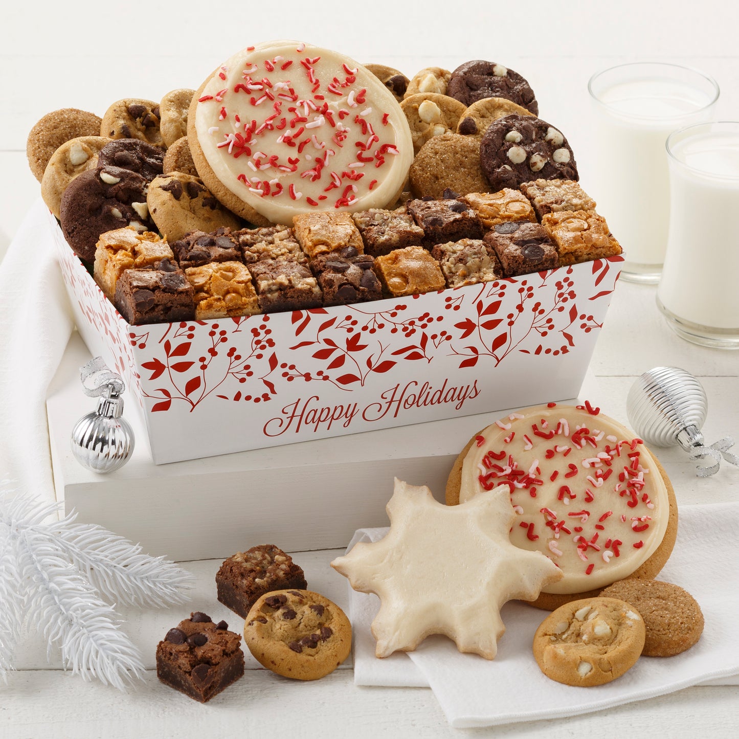 A Happy Holidays crate decorated with red holly leaves and berries and filled with an assortment of Nibblers®, brownie bites, two frosted peppermint cookies with sprinkles, and one frosted snowflake cookie. 