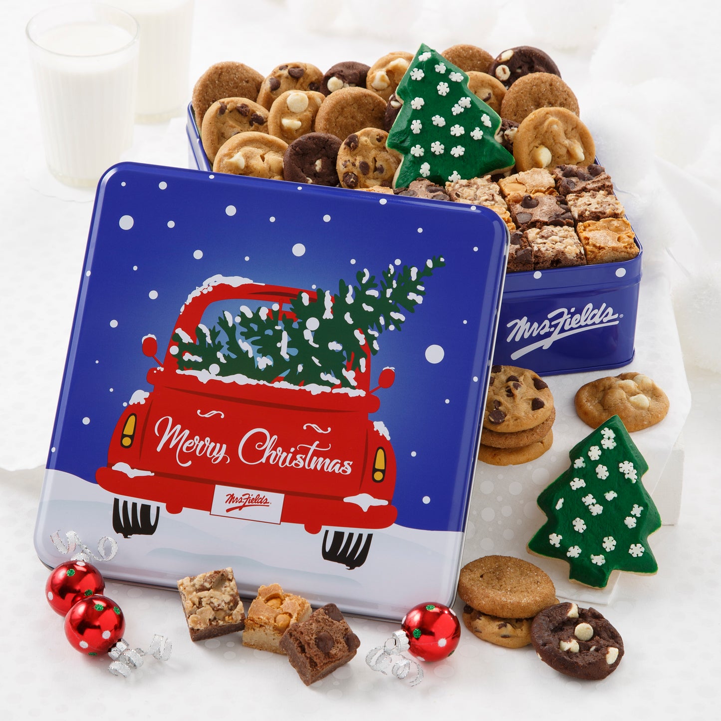 A blue Merry Christmas tin decorated with an old-fashioned red truck with a holiday tree in the back and filled with Nibblers®, brownie bites, and two frosted Christmas tree cookies.