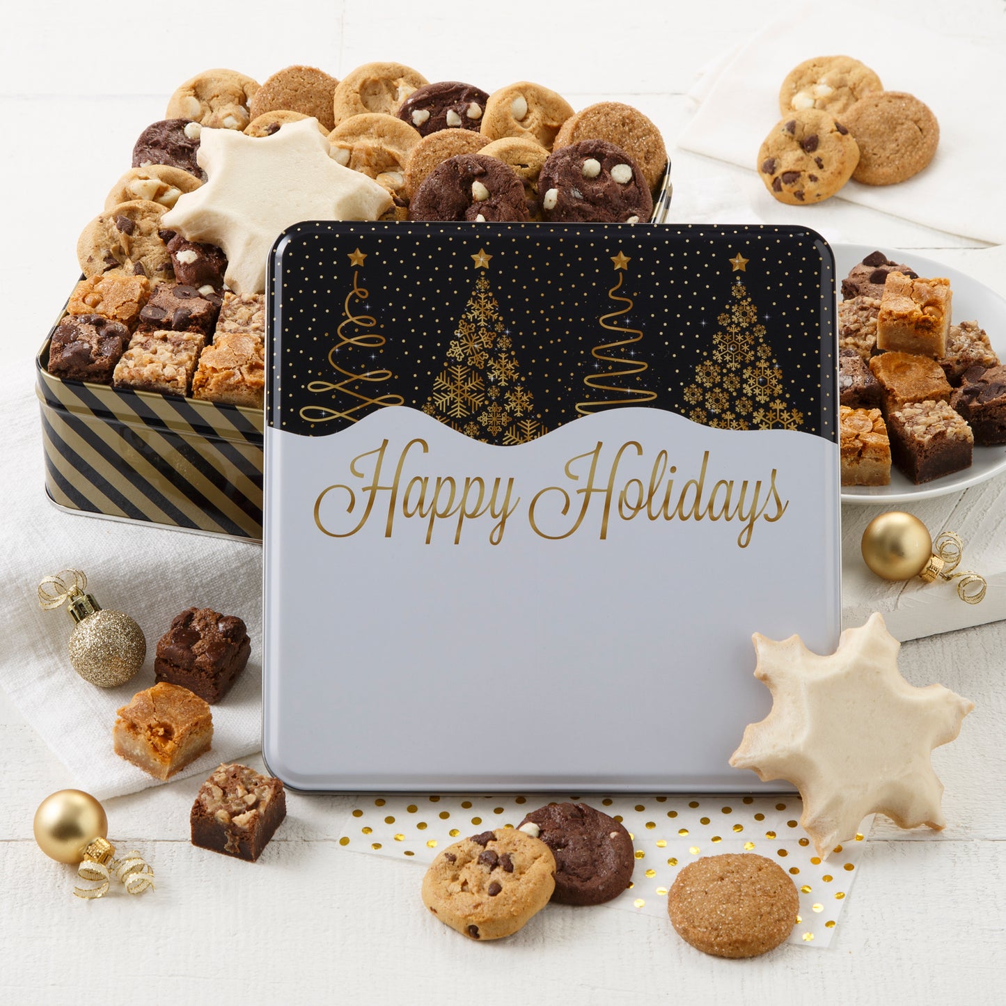 A happy holiday black and gold tin decorated with Christmas trees and filled with an assortment of Nibblers®, brownie bites, and two frosted snowflake cookies.