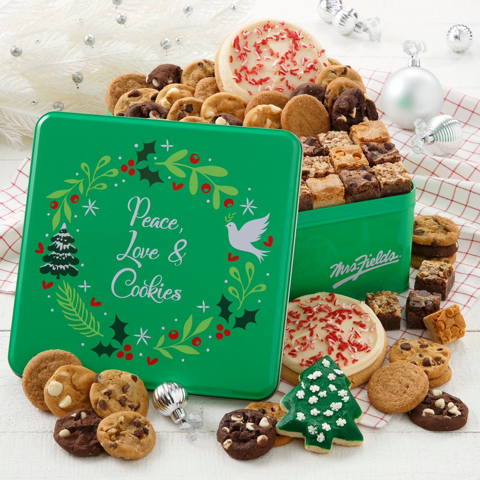 Green Peace, Love, & Cookies tin decorated with a holiday wreath and filled with an assortment of Nibblers®, brownie bites, two frosted peppermint round cookies with sprinkles and a frosted holiday tree cookie with sprinkles