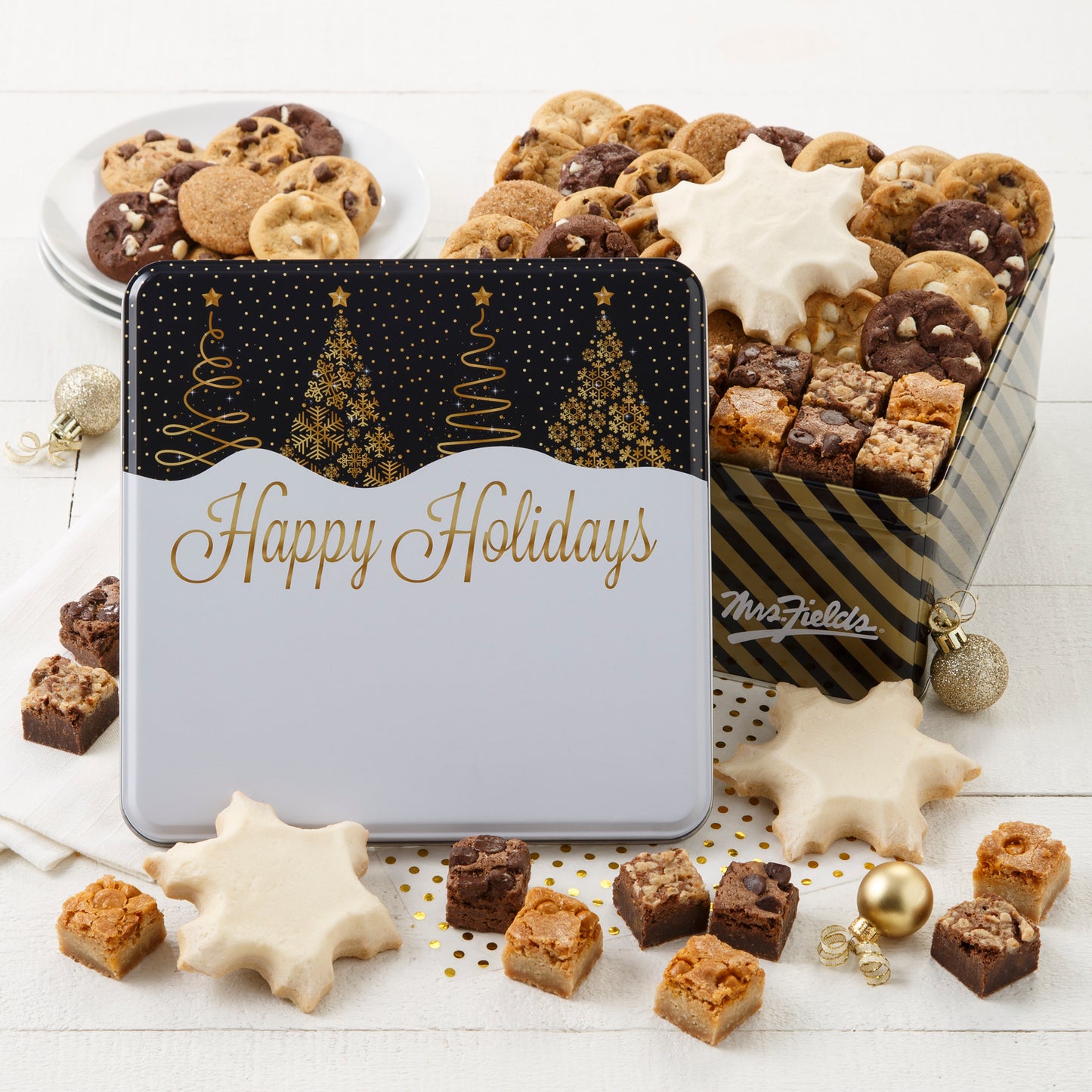 A Happy Holidays black and gold tin decorated with Christmas trees and filled with an assortment of Nibblers®, brown bites, and three frosted snowflakes.