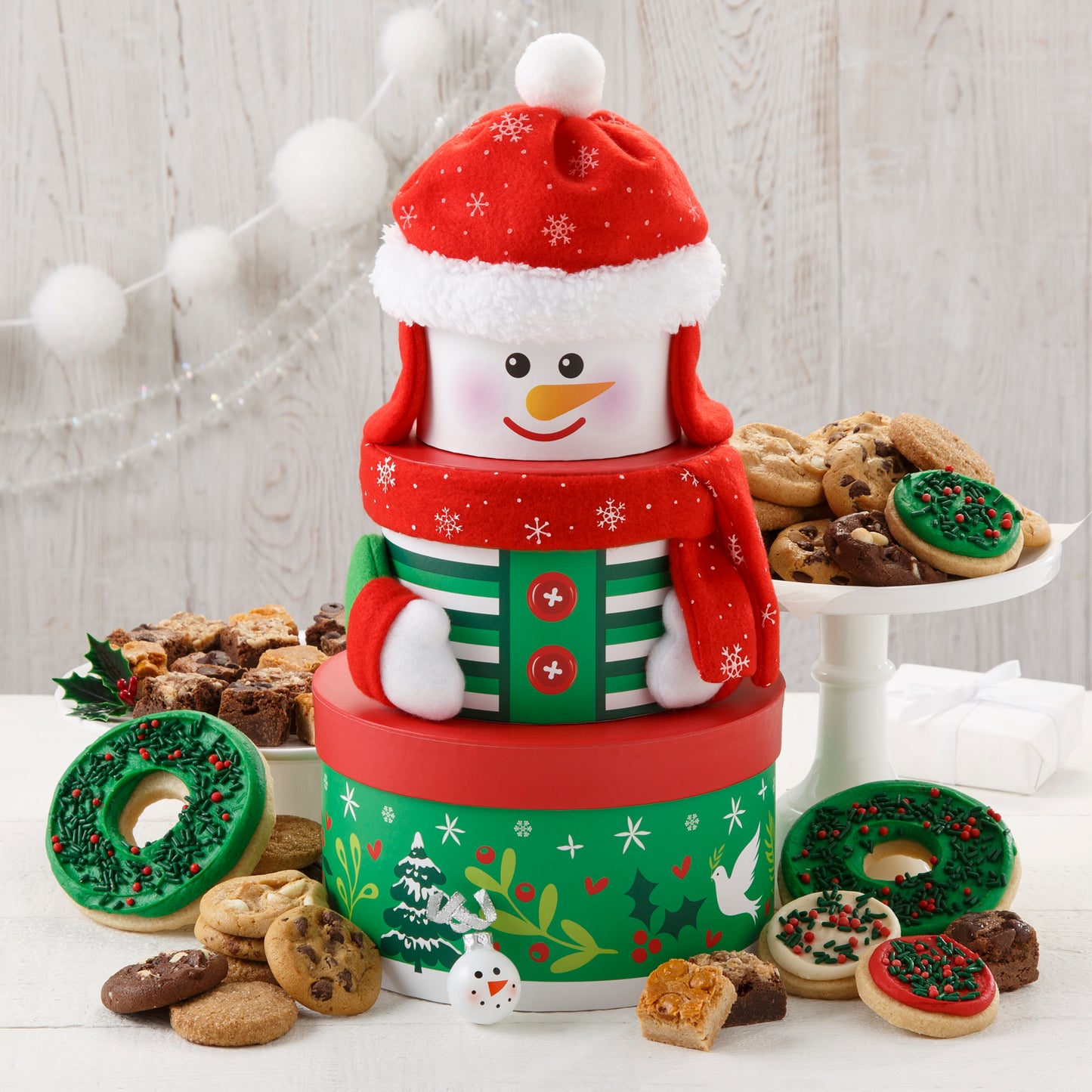 A green, red, and white snowman small tower that is surrounded by an assortment of Nibblers®, brownie bites, two frosted wreath cookies with sprinkles, and three frosted mini cookies with sprinkles. 