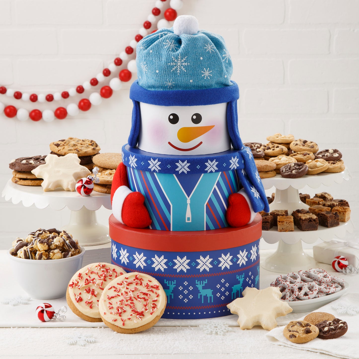 A blue, red, and white snowman tower surrounded by an assortment of Nibblers®, brownie bites, two frosted snowflakes, two frosted peppermint cookies with sprinkles, yogurt-covered pretzels, and chocolate drizzled popcorn.