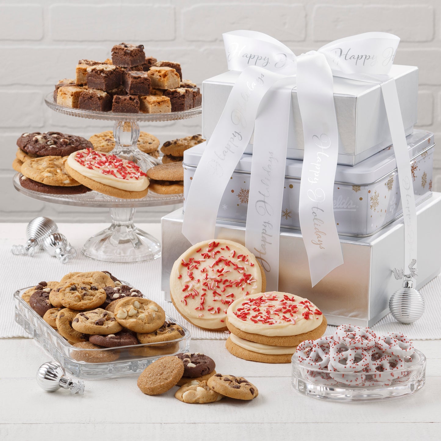 A white, silver, and gold three-tier tower that is surrounded by an assortment of Nibblers®, brownie bites, three frosted peppermint cookies, and yogurt-covered pretzels.