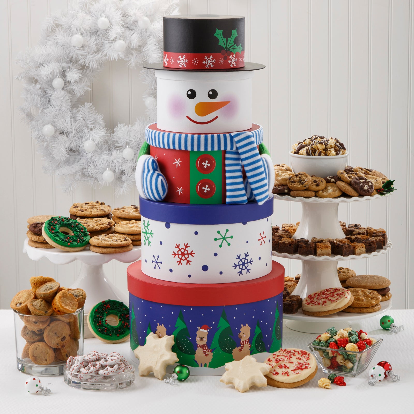 A festive five-tier snowman tower surrounded by an assortment of Nibblers®, original cookies, muffins, two frosted wreath cookies with sprinkles, two frosted peppermint cookies with sprinkles, two frosted snowflake cookies,  holiday popcorn, and yogurt-covered pretzels. 